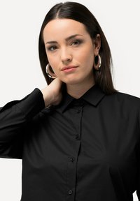 Camicia nera con bottoni realizzata in tessuto morbido; presenta un colletto e maniche lunghe con polsini abbottonati. Orecchini a cerchio argentati completano il look.