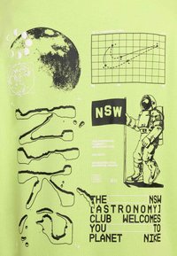 Grøn T-shirt med sorte grafiske designs, herunder en planet, astronaut, diagrammer og tekstelementer. Har iøjnefaldende, kontrasterende tryk.