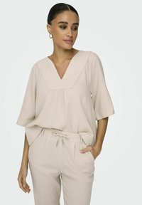Beige V-hals blouse met korte mouwen, met een geplooid voorkantontwerp. Draag het met bijpassende hoge taillebroek, ook in beige.