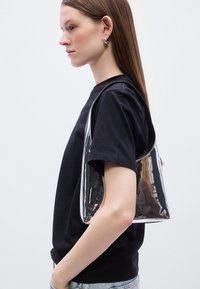 Ασημένια μεταλλική τσάντα ώμου με λεία υφή και στρογγυλεμένο σχήμα, φορεμένη στον ώμο, σε συνδυασμό με μαύρο μπλουζάκι και τζιν παντελόνια.