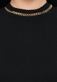 Maglione nero a coste con collo alto, caratterizzato da un accento di catena dorata attorno al collo. Tessuto ricco e dettaglio hardware lucido contrastante.