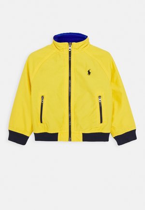 Gelbe Zip-up-Jacke mit schwarzen Bündchen und Saum, zwei Fronttaschen mit Reißverschluss und kleinem schwarzem Logo auf der Brust.