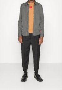 Graue Zip-Jacke über einem orangen Shirt, kombiniert mit schwarzen, taillierten Hosen und schwarzen Schuhen. Rotes Etikett-Detail auf der Jacke.