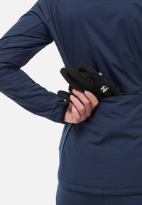 Des gants noirs sont tenus dans une main, contrastant avec un haut de sport bleu marine. Le tissu est lisse avec une coupe ajustée et des détails de couture subtils.