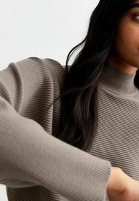 Sweter dzianinowy w odcieniu taupe z wysokim kołnierzem. Posiada teksturę ściągacza i długie rękawy. Zbliżenie na szczegóły ramienia i rękawa.