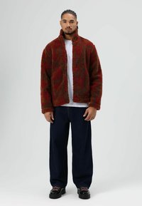 Vêtements : veste en peluche rouge et verte à motifs, col haut, fermeture éclair intégrale, associée à un jean large foncé et des chaussures noires.