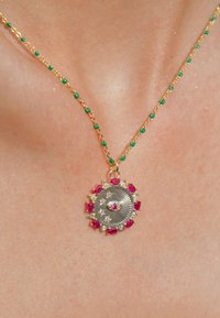 Collier en chaîne dorée avec des perles vertes ; présente un pendentif circulaire avec un design d'œil central, entouré de pierres précieuses roses et transparentes.
