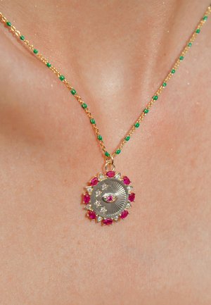 Collier en chaîne dorée avec des perles vertes ; présente un pendentif circulaire avec un design d'œil central, entouré de pierres précieuses roses et transparentes.