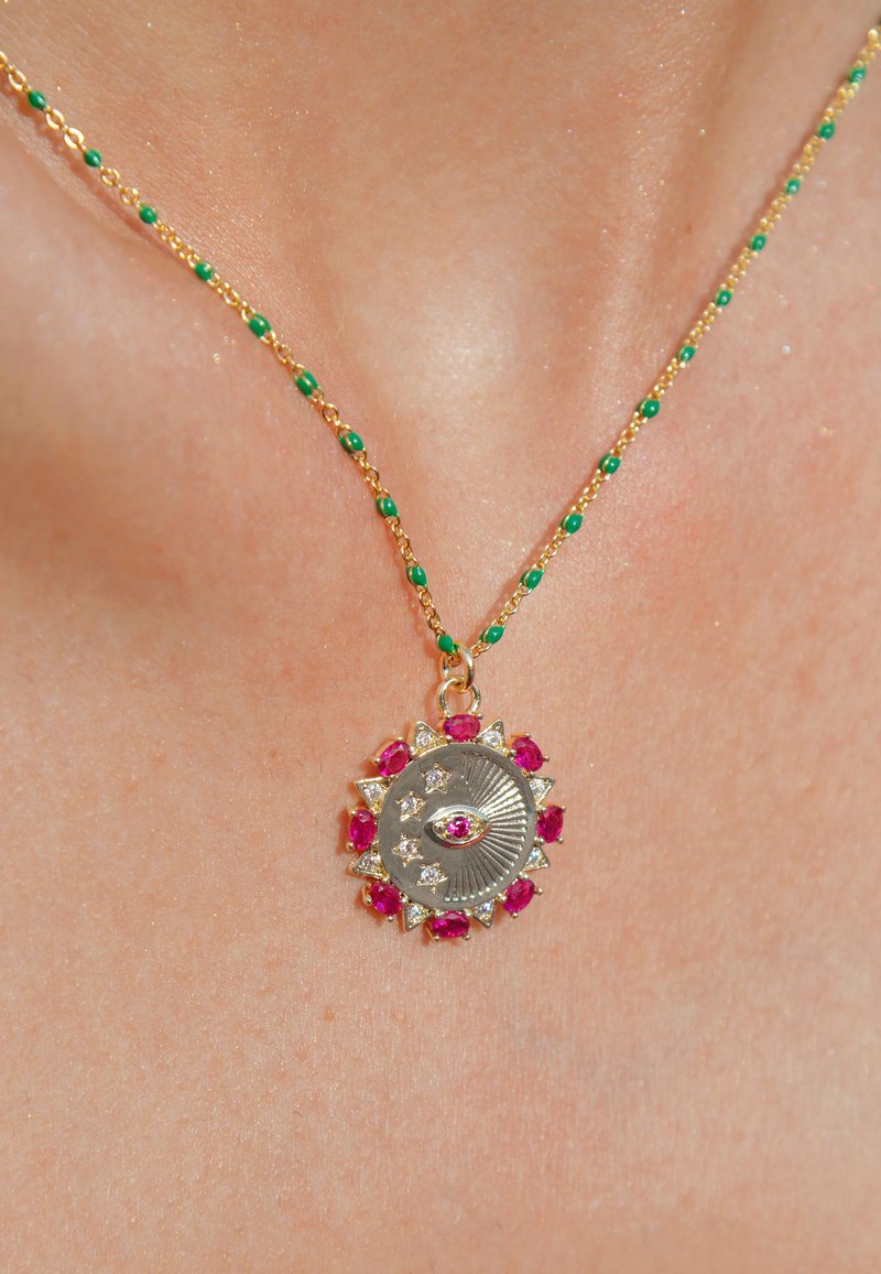 Collier en chaîne dorée avec des perles vertes ; présente un pendentif circulaire avec un design d'œil central, entouré de pierres précieuses roses et transparentes.
