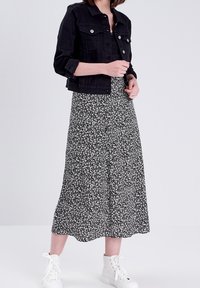 Zwarte spijkerjack met zilverkleurige hardware, gelaagd over een zwarte maxi-jurk met bloemenprint. Model draagt witte enkelboots.