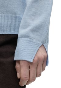 Χέρι μερικώς καλυμμένο από μανσέτα γαλάζιου πουλόβερ με άνοιγμα για τον αντίχειρα, ακουμπισμένο σε σκούρο καφέ παντελόνι.