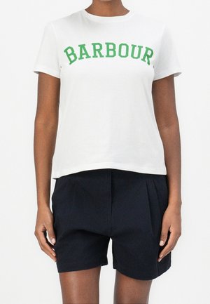 Biała koszulka z krótkim rękawem z zielonym napisem "BARBOUR" połączona z czarnymi plisowanymi szortami z wysokim stanem na osobie na białym tle.