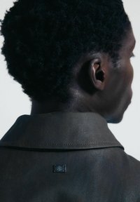 Giubbotto in pelle marrone con una texture sottile, caratterizzato da una piccola placca metallica con logo all'altezza del colletto. Il colletto è leggermente rialzato.