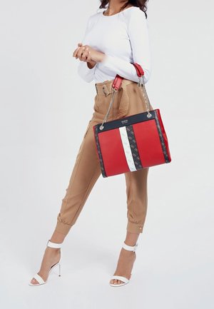 Mujer con pantalones beige y blusa blanca sosteniendo un bolso grande rojo, negro y blanco con correas de cadena, lleva sandalias blancas de tacón alto.