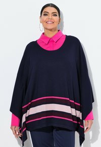 Frau mit Creolen, hellrosa Hemd mit Kragen unter einem marineblauen Poncho mit rosa, burgunderrot und beige gestreift, lächelnd vor einer weißen Wand.