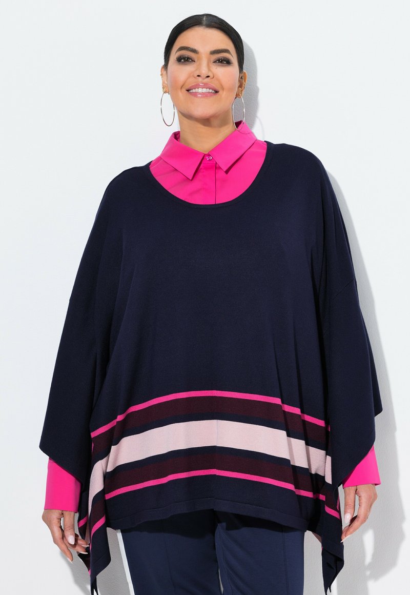 Frau mit Creolen, hellrosa Hemd mit Kragen unter einem marineblauen Poncho mit rosa, burgunderrot und beige gestreift, lächelnd vor einer weißen Wand.