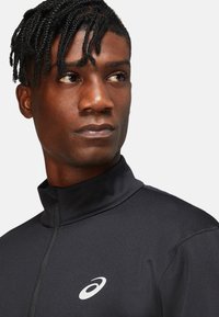 Black athletic jacket with a high collar, zip closure, and silver logo. Smooth texture and fitted design suitable for performance wear.