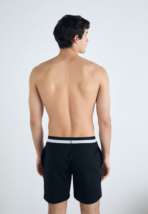 Maillot de bain noir avec une ceinture blanche contrastante, à la texture lisse et doté de poches latérales, vu de l'arrière.
