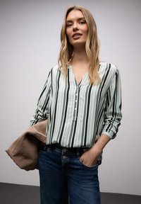 Femme aux cheveux blonds longs portant un chemisier à rayures verticales vertes et blanches et un jean bleu, tenant une veste beige sur un bras.