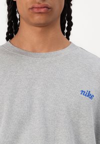 Sweatshirt cinza em algodão com gola redonda, apresentando um logótipo "nike" bordado em azul no lado esquerdo do peito. Textura suave e corte relaxado.