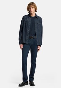 Dunkelblaue Jeanshemd mit zwei Brusttaschen, getragen über einem marineblauen T-Shirt. Kombiniert mit dunkelblauen Hosen und schwarzen Schuhen.
