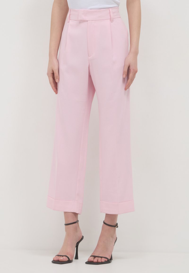 Donna che indossa pantaloni a vita alta larghi rosa chiaro e sandali con tacchi alti e cinturini neri, in piedi davanti a uno sfondo semplice.