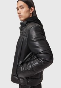 Sort læder pufferjakke med lynlås foran, ribbede manchetter, høj krave og en struktureret sort fleecefoer nær hætten.