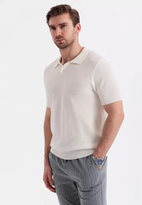 Krémová polo košile s krátkým rukávem a texturovaným vzorem, s dvoupoložkovým límcem a žebrovaným lemem. V kombinaci s šedými kalhotami s proužkem.