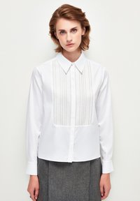 Blusa blanca de manga larga con cuello abotonado, que presenta pliegues verticales texturizados en el frente y una tela suave en todo el resto.