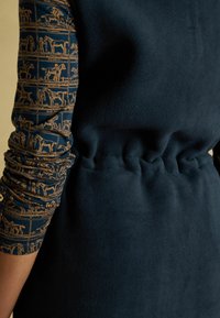 Navy fleece beklædning med rynket talje ved siden af armen i blå ærme med mønster af gyldne heste og ryttere i rektangulære rammer.