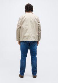 Άνδρας με κοντά σγουρά μαλλιά, φοράει μπεζ μπουφάν, μπλε τζιν και καφέ παπούτσια, στέκεται με την πλάτη σε απλό φόντο.