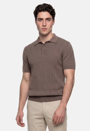 Junger Mann mit kurzen lockigen Haaren, der ein braunes kurzärmeliges Pullunder-Poloshirt und beige Hosen trägt, steht vor einem schlichten Hintergrund.