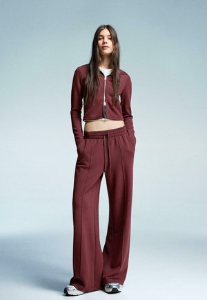 Młoda kobieta o długich włosach, ubrana w bordową, zapinaną na zamek kurtkę skróconą i pasujące szerokie spodnie oraz tenisówki, z rękami w kieszeniach.