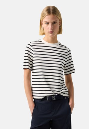 Kvinde med kort blondt hår iført en hvid og sort stribet kortærmet skjorte og mørke bukser med hænderne i lommerne.