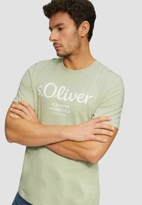 Light green cotton t-shirt featuring white printed text, crew neckline, and short sleeves. Model poses with arms crossed.