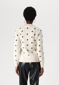 Pull en tricot crème avec des motifs d'étoiles noires, poignets et ourlet côtelés, col rond, texture douce, associé à un pantalon en cuir noir.