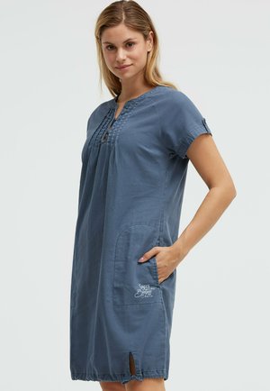 Frau trägt ein knielanges, kurzärmeliges blaues Kleid mit Falten, einer Fronttasche und kleinem Bindedetail am Saum und steht mit einer Hand in der Tasche.