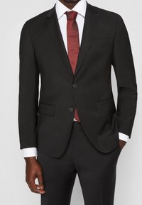 Zwarte op maat gemaakte colbertsjas met twee voorzakken, sluiting met één knoop en een rode stropdas. Witte shirtmanchetten zichtbaar aan de mouwen.