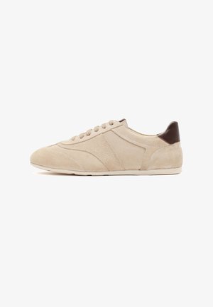 Beige suède lage sneaker met bruine leren hielstuk, ronde neus en vetersluiting op een witte achtergrond.