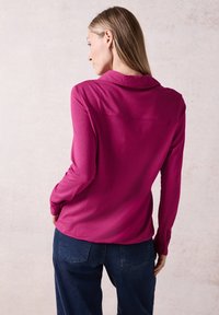 Långärmad, kragskjorta i klart fuchsia-tyg, med ryggslå och slät textur, kombinerad med mörkblå denimbyxor.