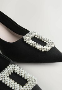 Ballerines en daim noir à bout pointu, ornées d'une grande boucle en rhinestones sur le devant, ajoutant un détail texturé à l'ensemble du design.