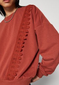 Mikina v barvě rezavé s texturovaným krajkovým detailem na levé straně, s žebrovanými manžetami a lemem pro těsné přilnutí.