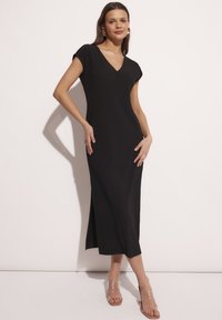 Czarna dzianinowa sukienka prążkowana z dekoltem w kształcie litery V i krótkimi rękawami, z bocznym rozcięciem. Noszona z przezroczystymi sandałami na obcasie, o gładkiej fakturze.