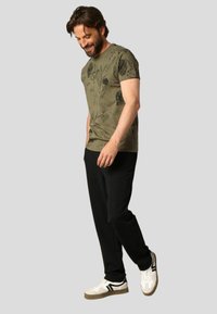 T-shirt vert olive avec motif floral, associé à un pantalon de jogging noir et des baskets blanches avec des accents noirs. Le mannequin sourit.