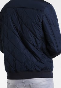 Marinblå quiltad jacka med diamantstygn, som bärs över ljusgrå jeans, som visar baksidan och sidorna.