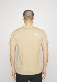 Béžové bavlněné tričko s krátkým rukávem, které má malé bílé logo na horní části zad. Jednoduchý, klasický design s kulatým výstřihem.