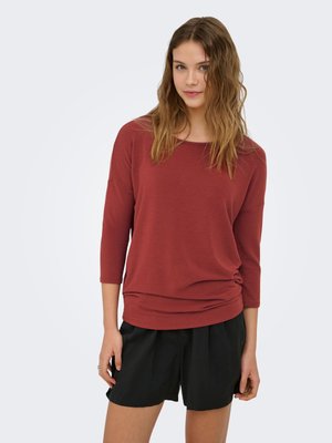 Jonge vrouw met golvend haar die een roestkleurige longsleeve top en zwarte geplooide shorts draagt, staand tegen een effen lichte achtergrond.