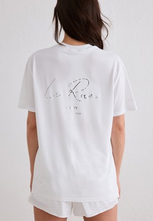 Kobieta o długich brązowych włosach, ubrana w białą, oversize'ową koszulkę z delikatnym napisem na plecach oraz białe spodenki, stojąca na tle jednolitej ściany.