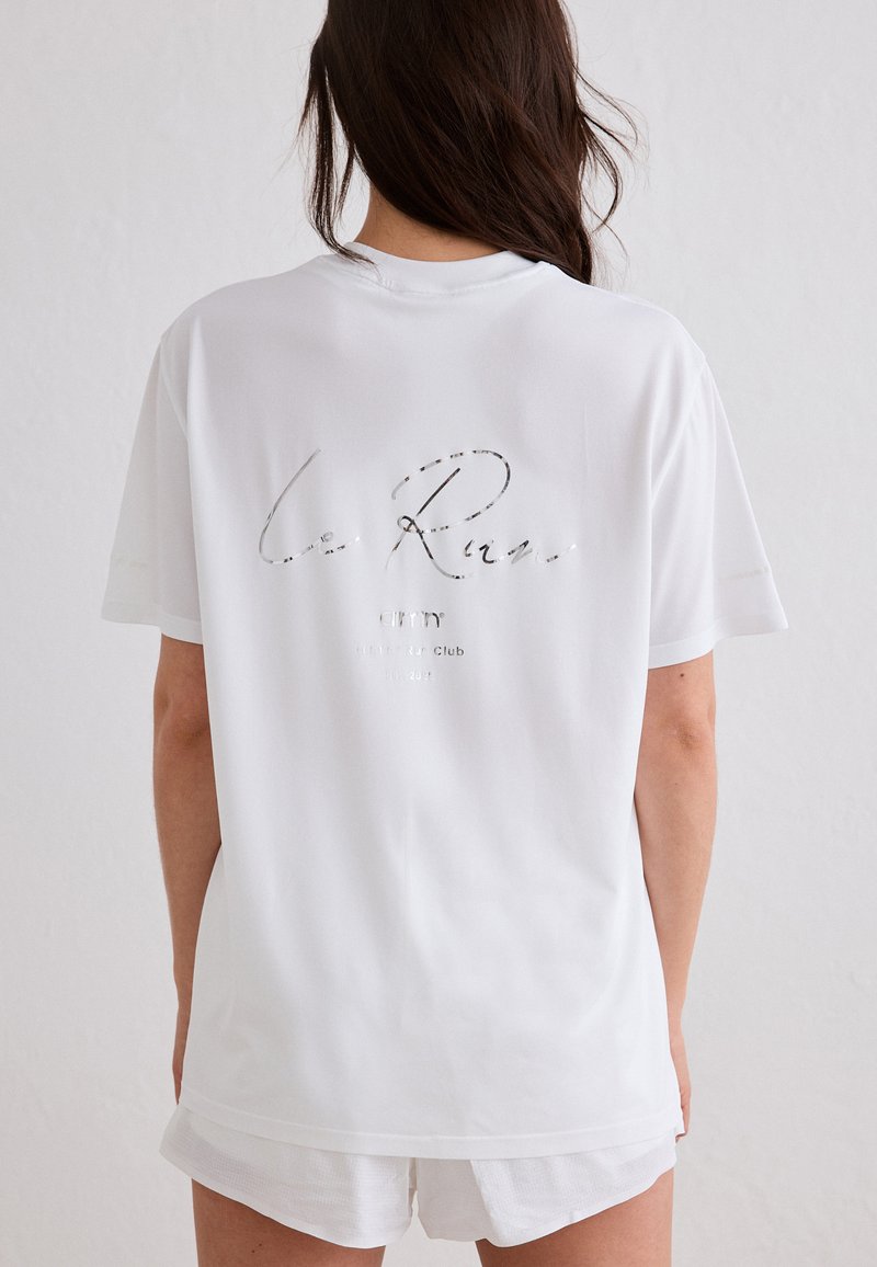 Femme aux longs cheveux bruns portant un T-shirt blanc oversize avec un texte léger au dos et un short blanc, debout contre un mur uni.