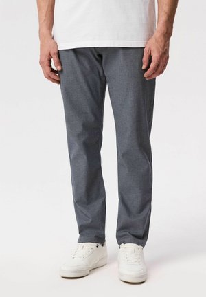 Mężczyzna w szarych prostych spodniach, białych trampkach i białej koszuli, stojący na jednolitym jasnym tle.